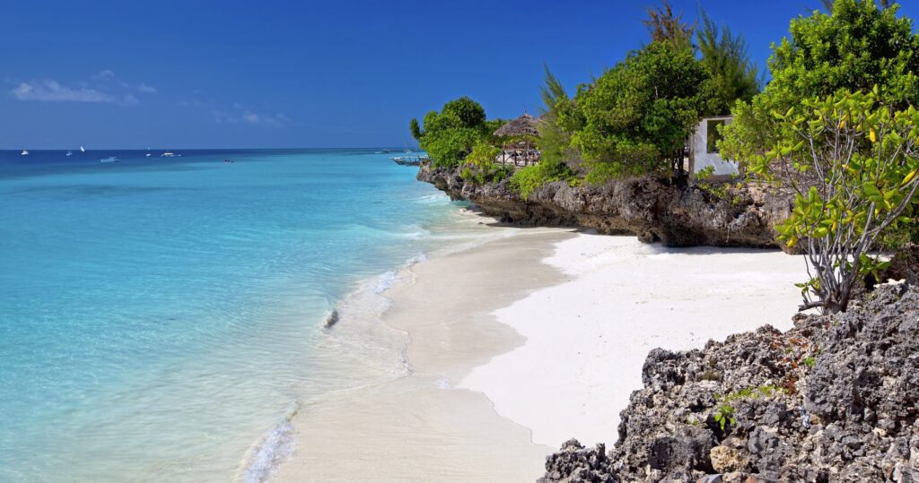 Blaues Meer mit weißem Strand vor braunen Felsen mit grünen Bäumen