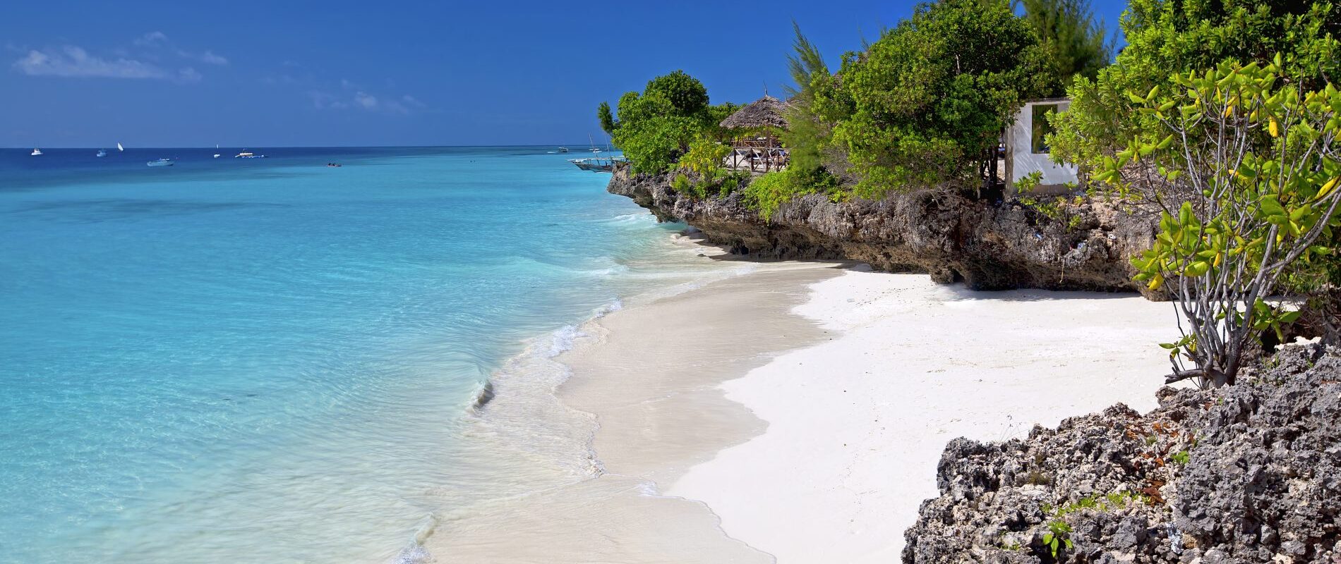 Blaues Meer mit weißem Strand vor braunen Felsen mit grünen Bäumen